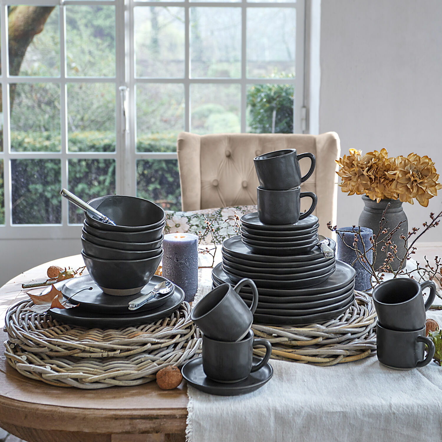 Černý keramický jídelní set na proutěných podložkách na dřevěném stole s dekoracemi u okna