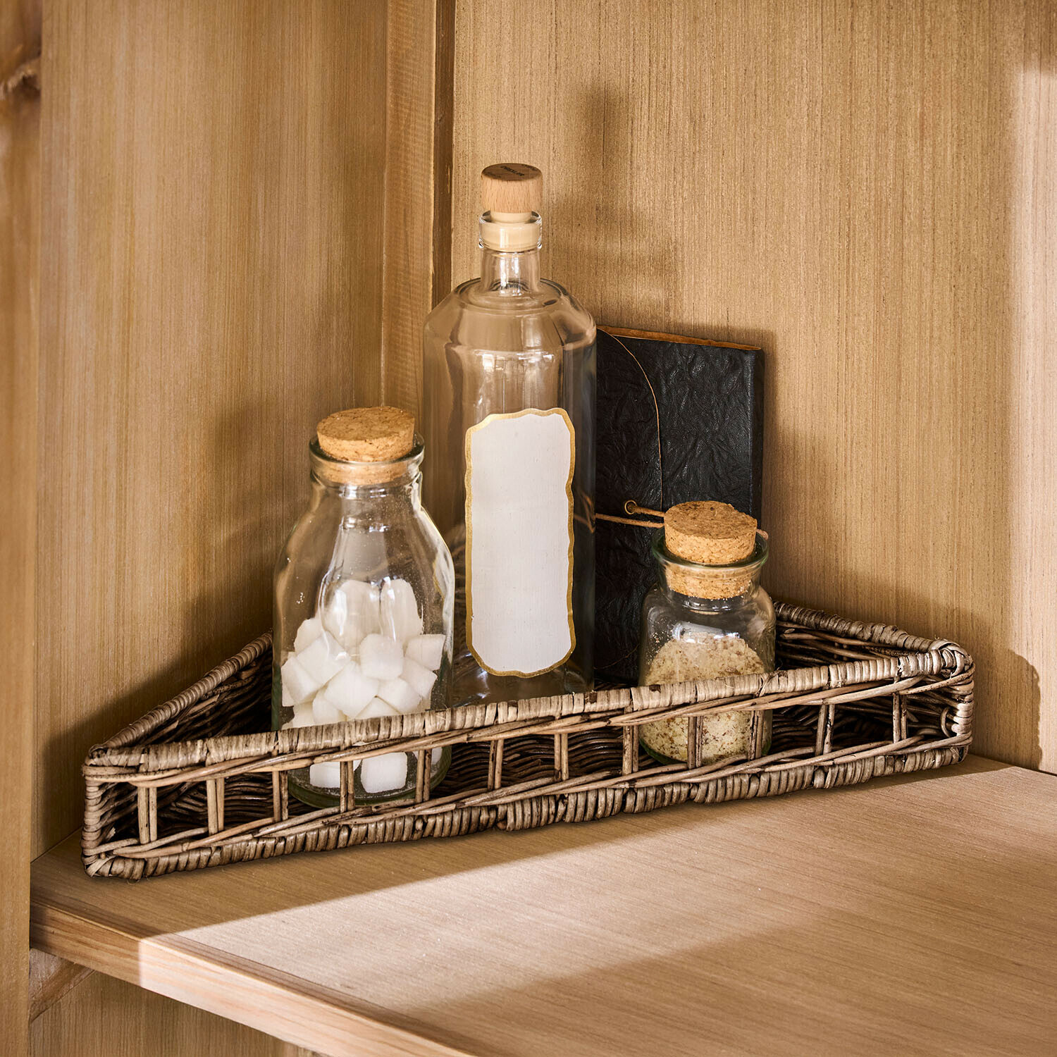 Trojúhelníkový ratanový košík v přírodní barvě s třemi skleněnými lahvemi a zápisníkem na dřevěné polici