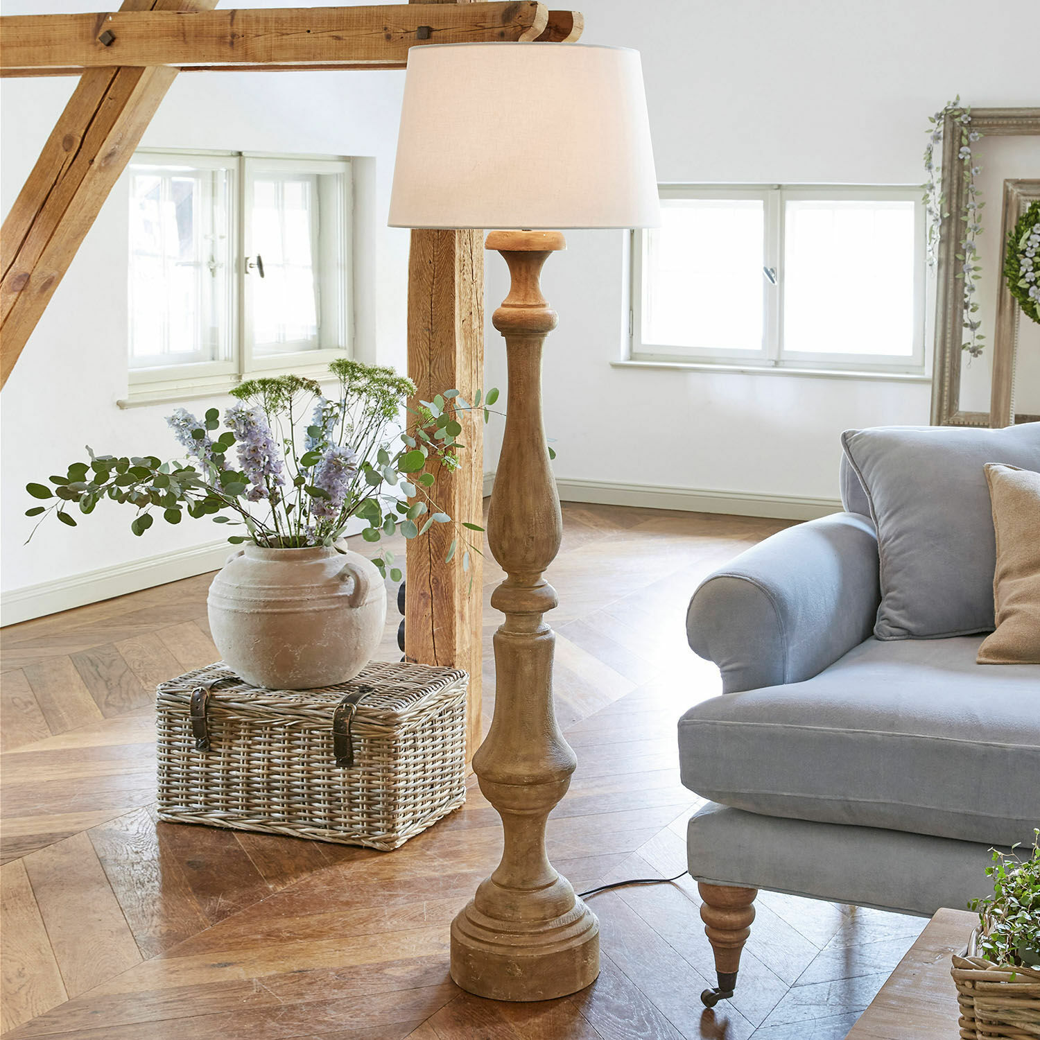Stojací lampa Etienne Dřevěná stojací lampa s bílým stínidlem v světlém obývacím pokoji s modrou pohovkou a proutěným košem