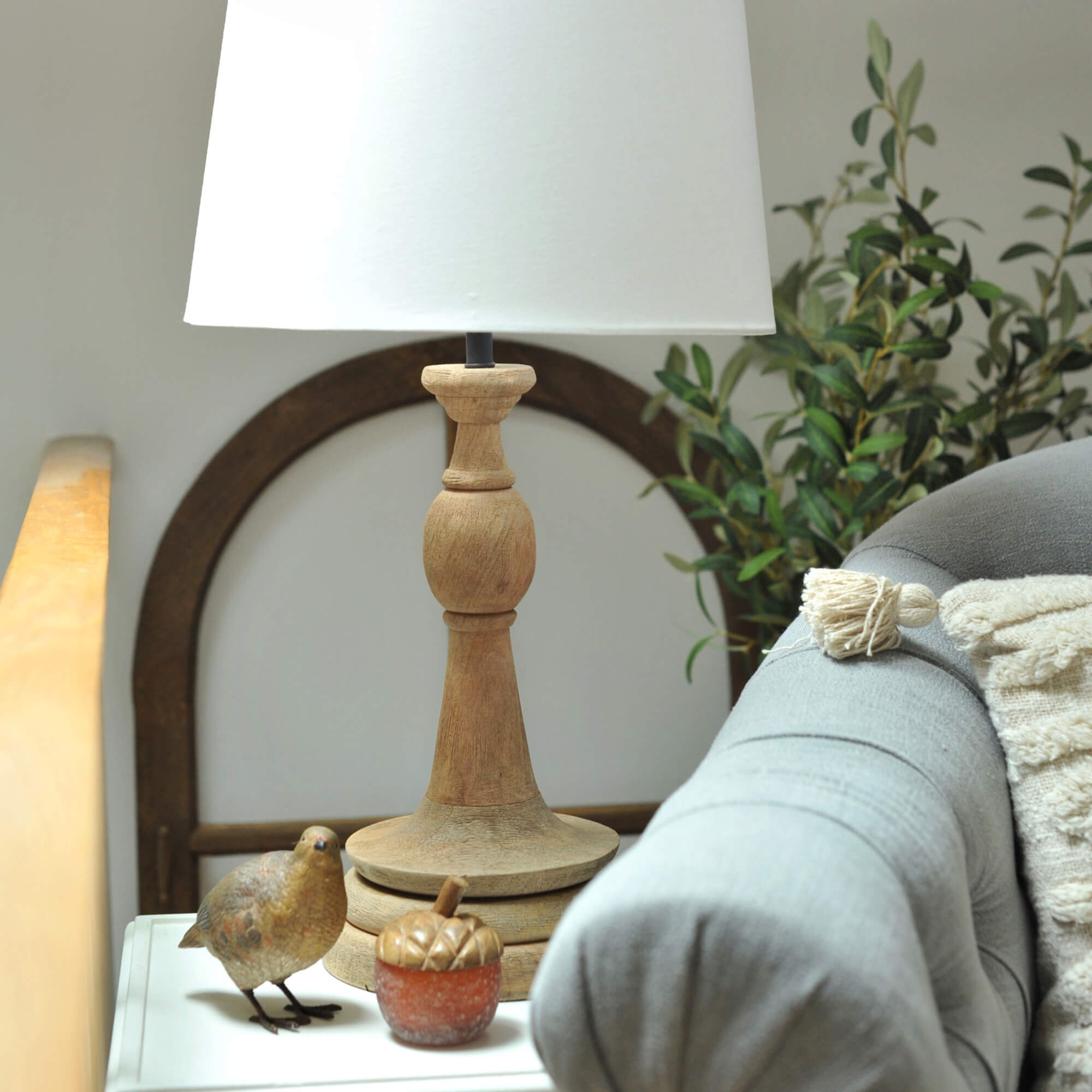 Dřevěná stolní lampa s bílým stínidlem na stolku vedle šedé pohovky a dekorace ptáka