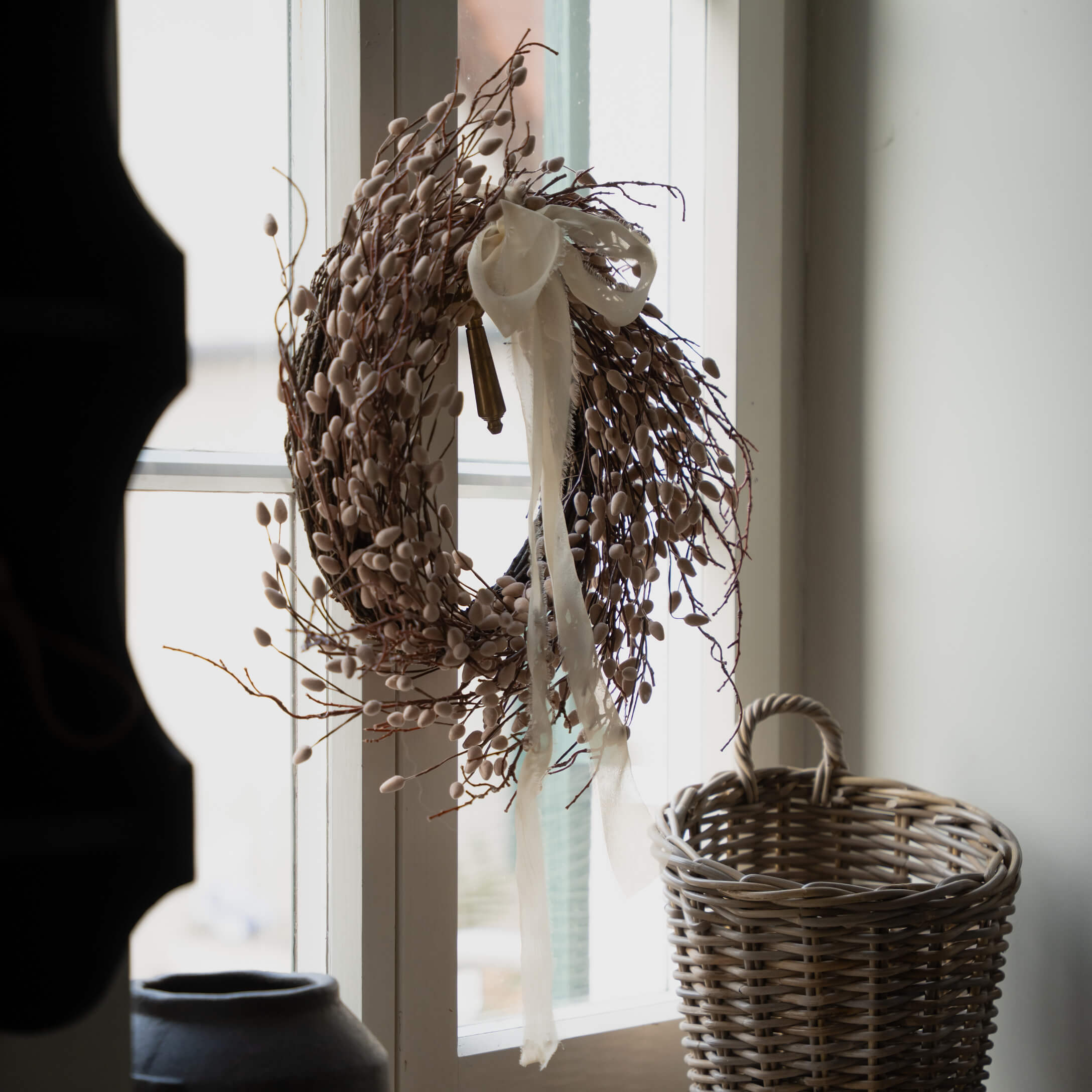 Dekorativní věnec z větviček a pupenů v béžové barvě s bílou stuhou na okně, vedle proutěný koš