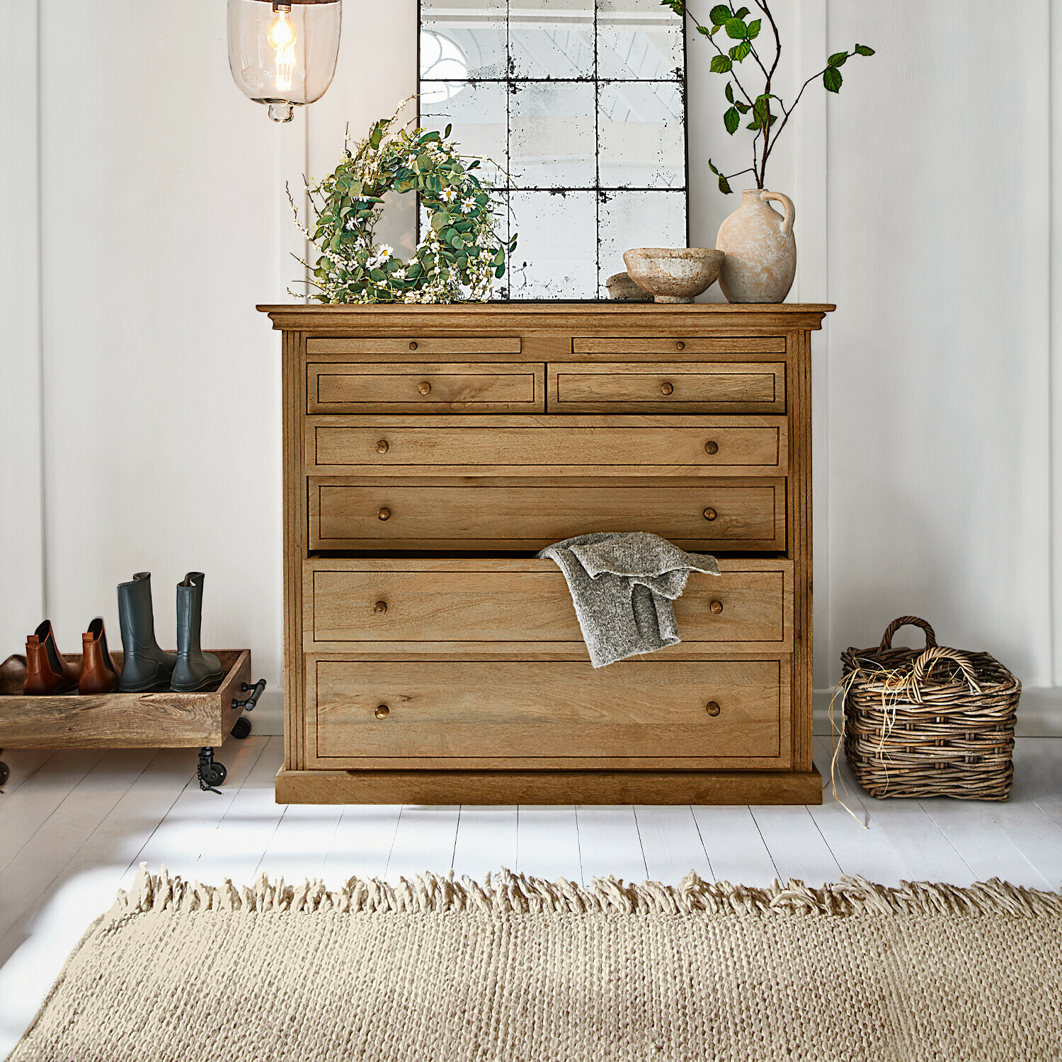 Dřevěná komoda v přírodní barvě s několika zásuvkami, dekoracemi a světlým kobercem v interiéru