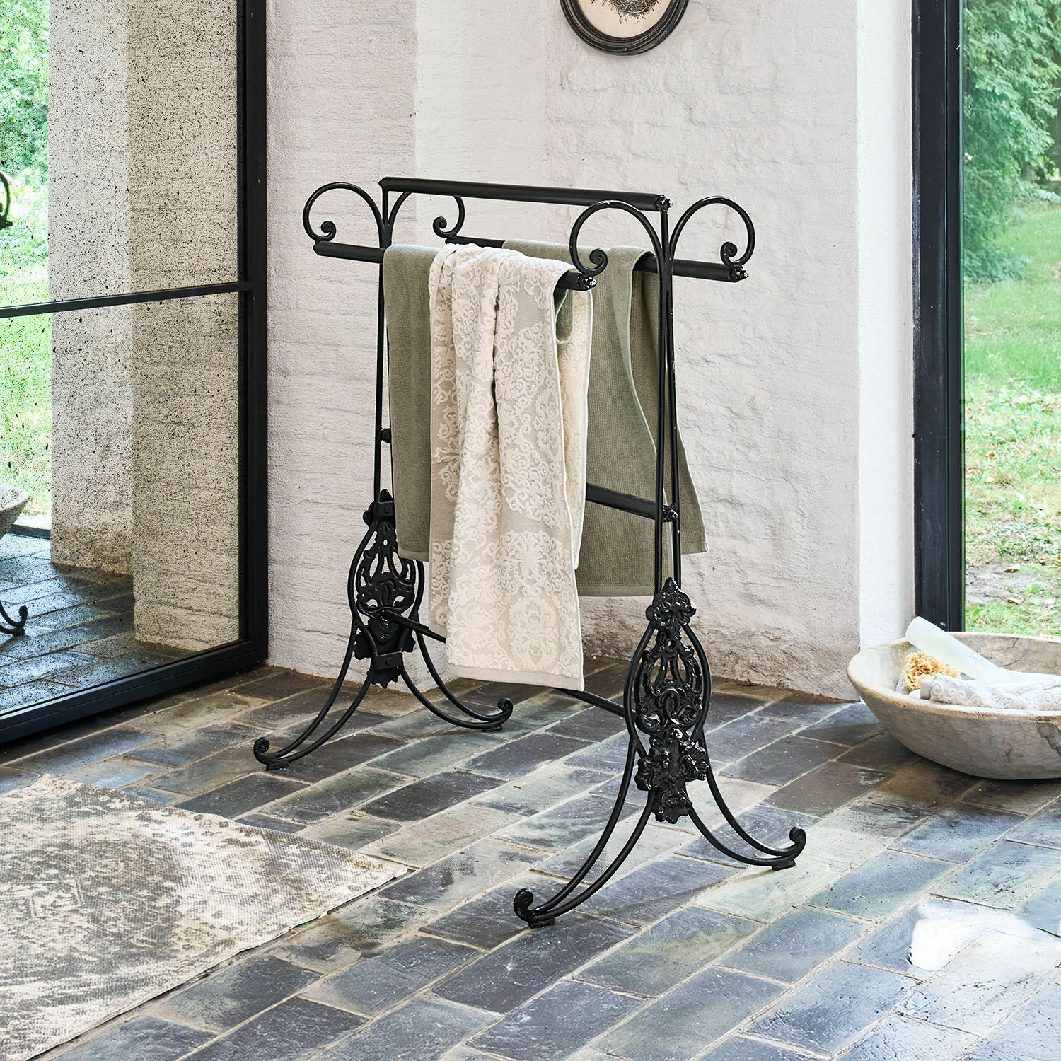 Černý kovový stojan na ručníky s ozdobnými detaily na kamenné podlaze v světlém interiéru
