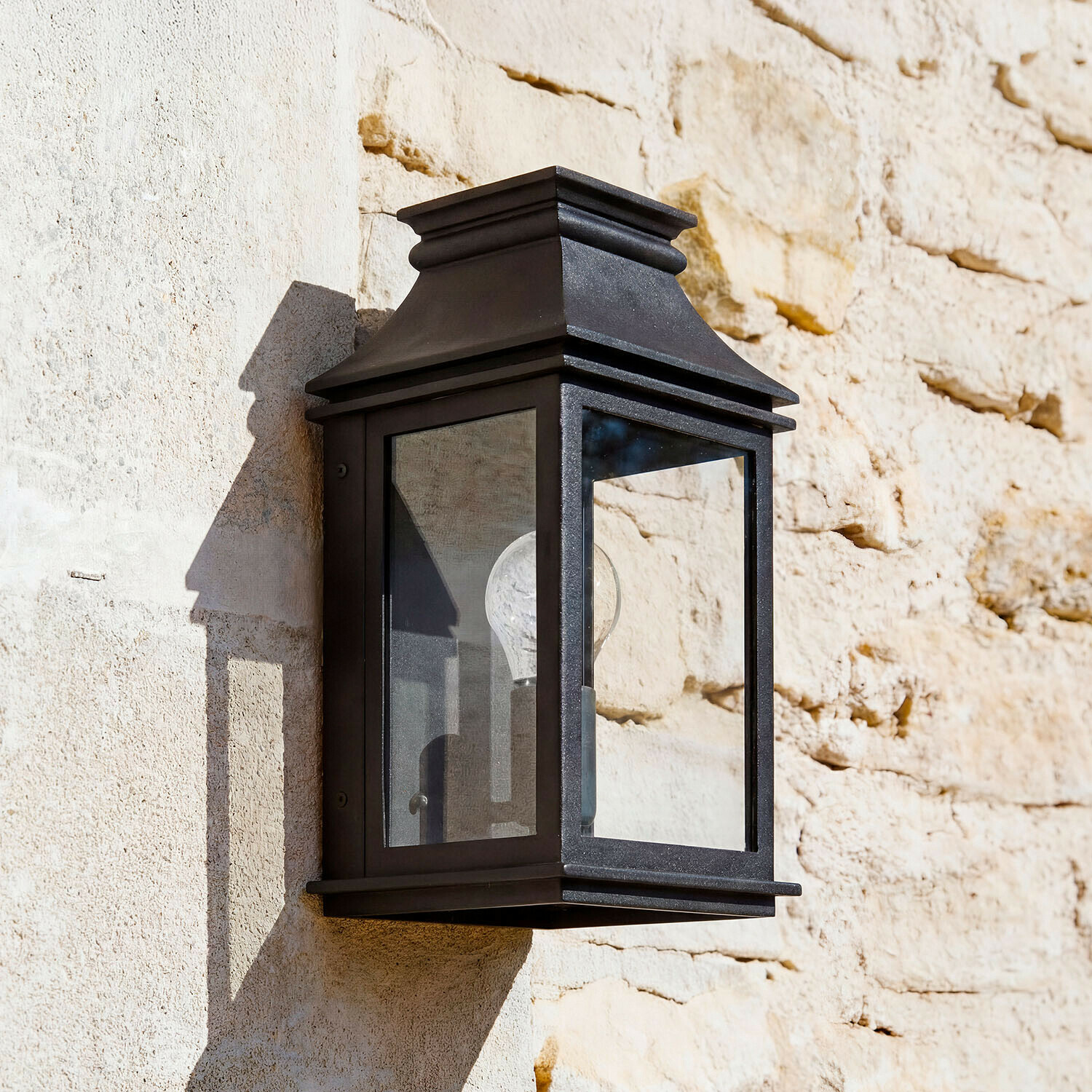 Venkovní nástěnná lampa Zélivarne Černé kovové nástěnné svítidlo s průhledným sklem na světlé kamenné zdi, venkovní osvětlení