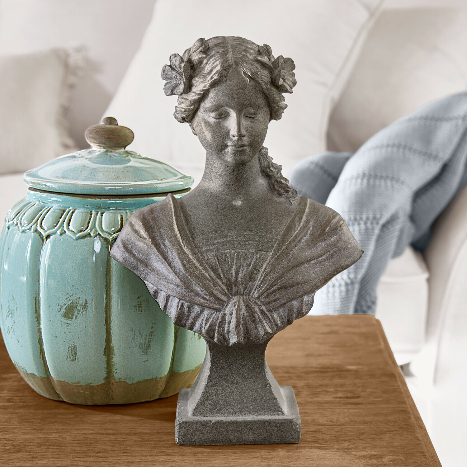 Šedý kamenný bust ženské postavy s květinami ve vlasech na dřevěném stole vedle tyrkysové keramické nádoby