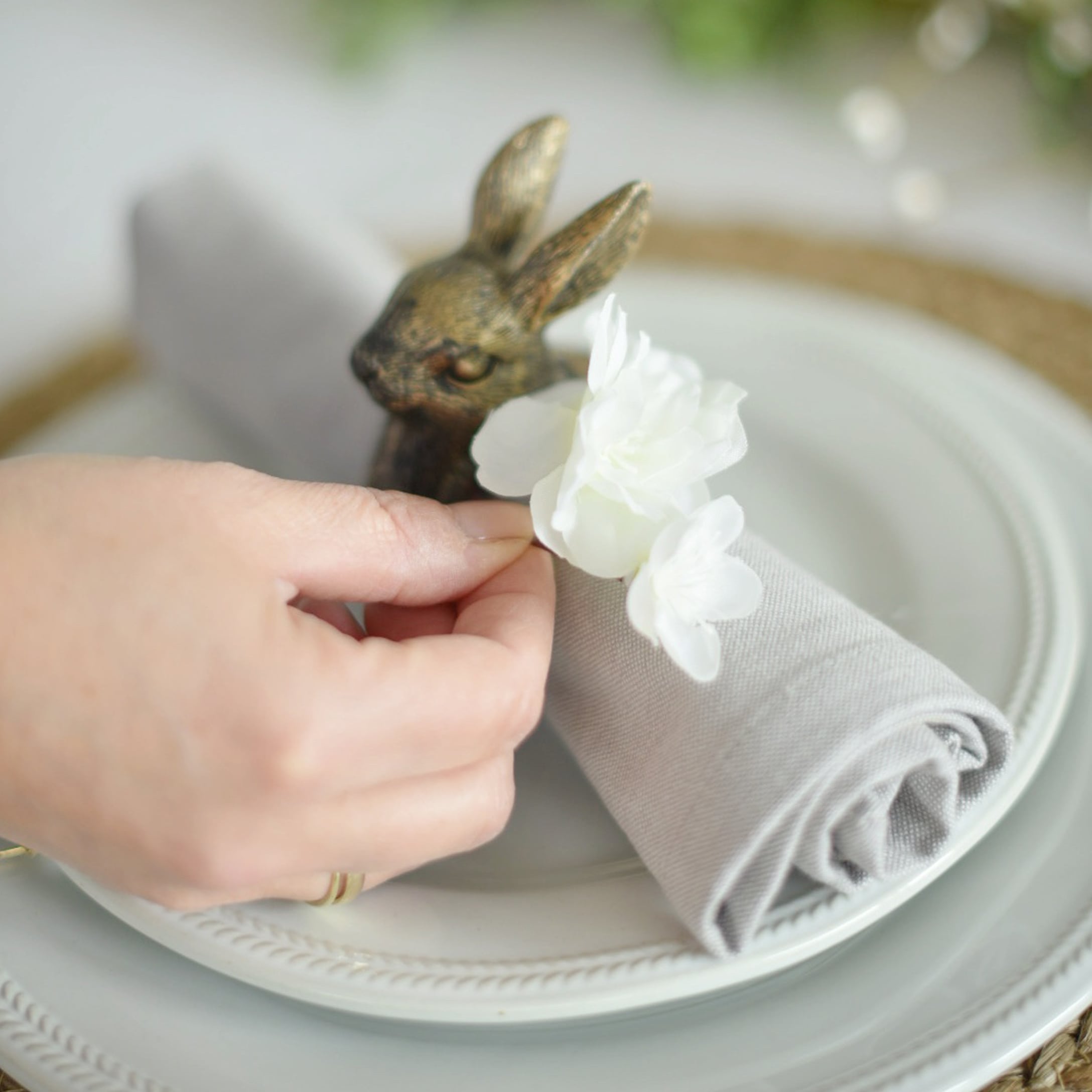 Šedý lněný kroužek na ubrousek s bílým květem a mosaznou dekorací zajíce na talíři