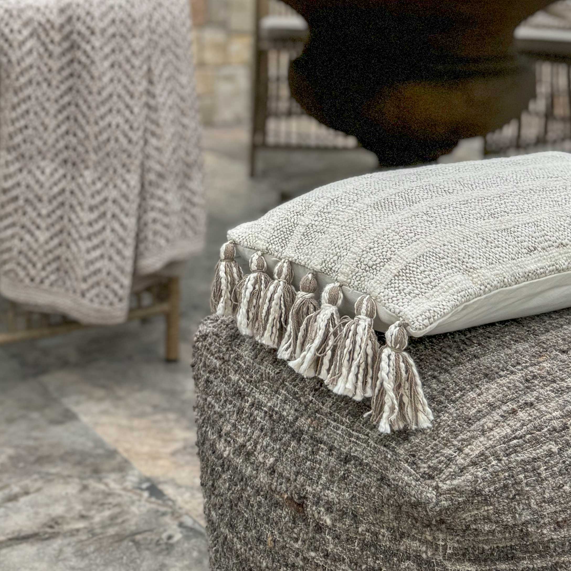 Béžový dekorativní polštář s třásněmi na šedém texturovaném pufu v boho stylu