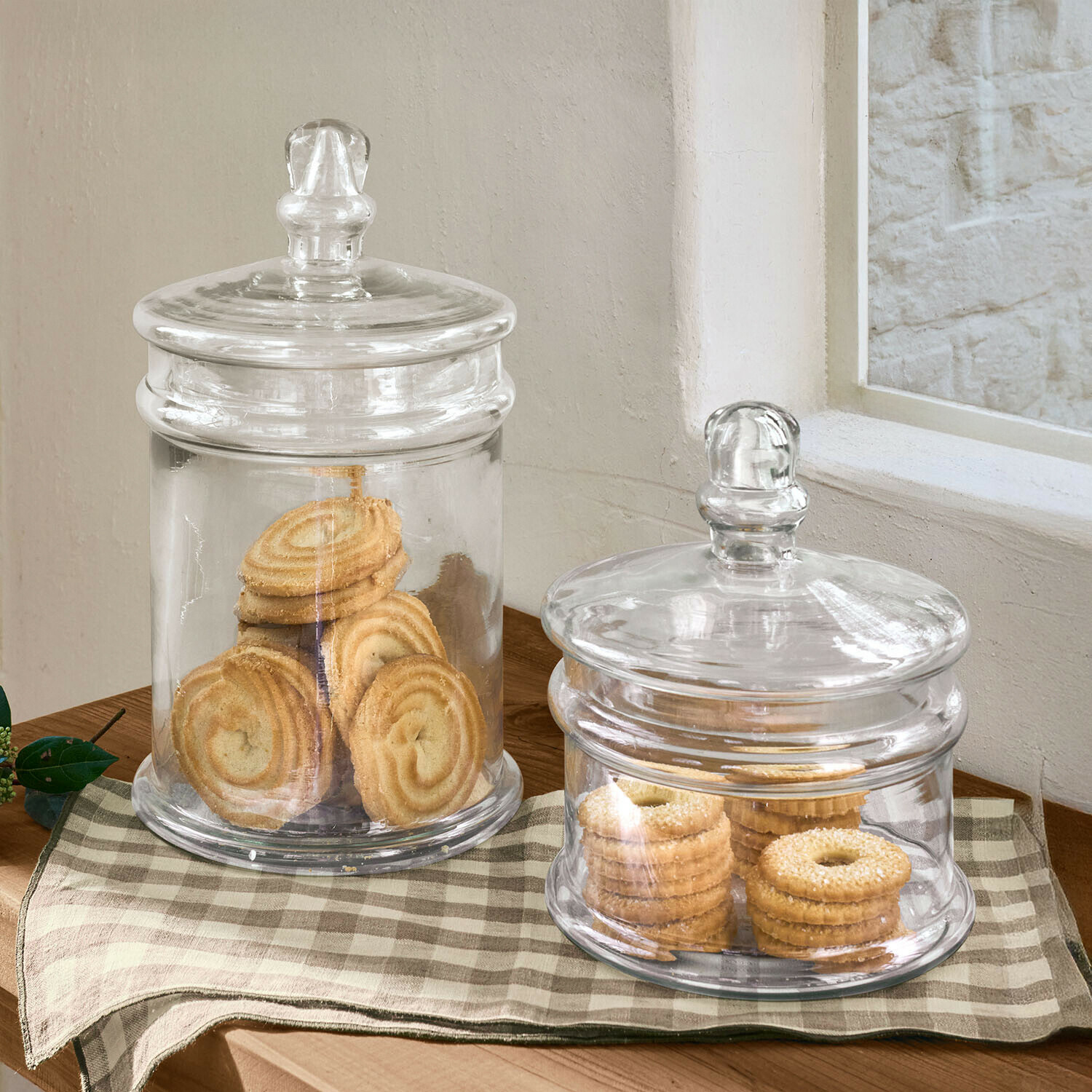 Dvě průhledné skleněné dózy s víčky plné sušenek na dřevěném stole s kostkovaným ubrusem