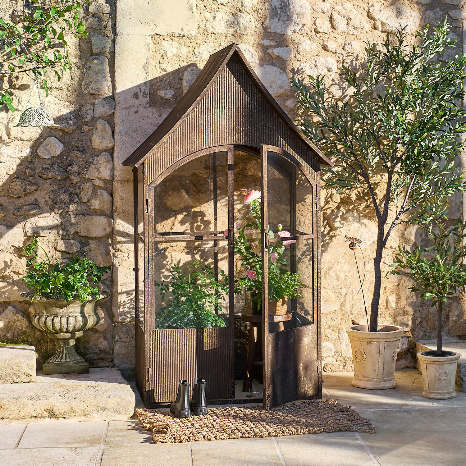 Zahradní kovová skleník ve vintage stylu hnědé barvy s rostlinami a květináči před kamennou zdí