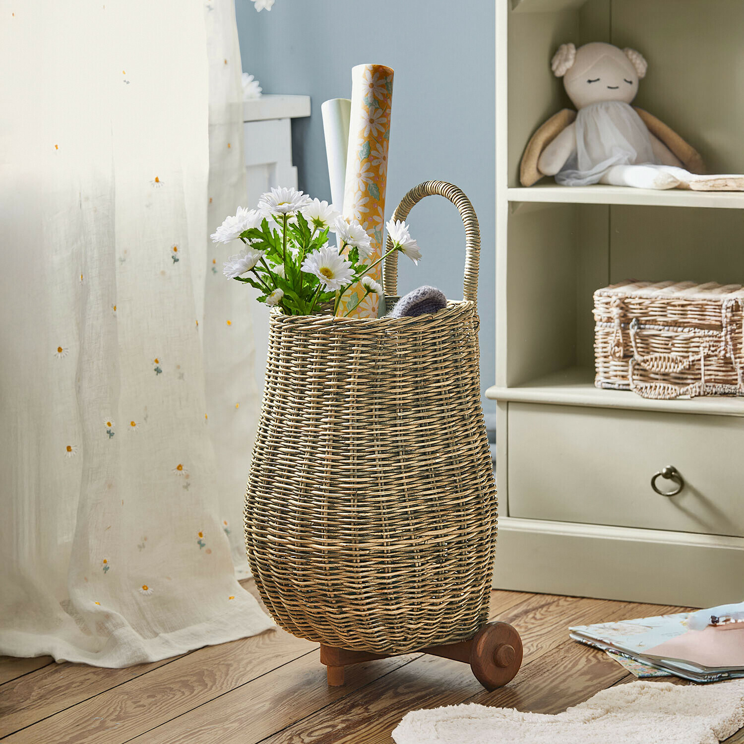 Přírodní proutěný koš na kolečkách s rukojetí, naplněný bílými květinami a dekoračním papírem