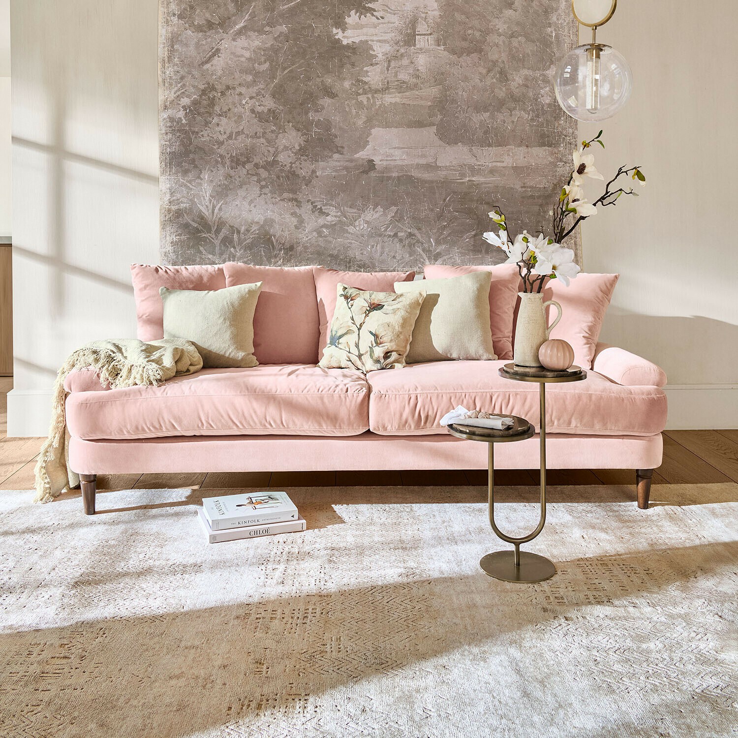 Růžová sametová pohovka s polštáři a odkládacím stolkem v světlém obývacím pokoji s nástěnnou dekorací