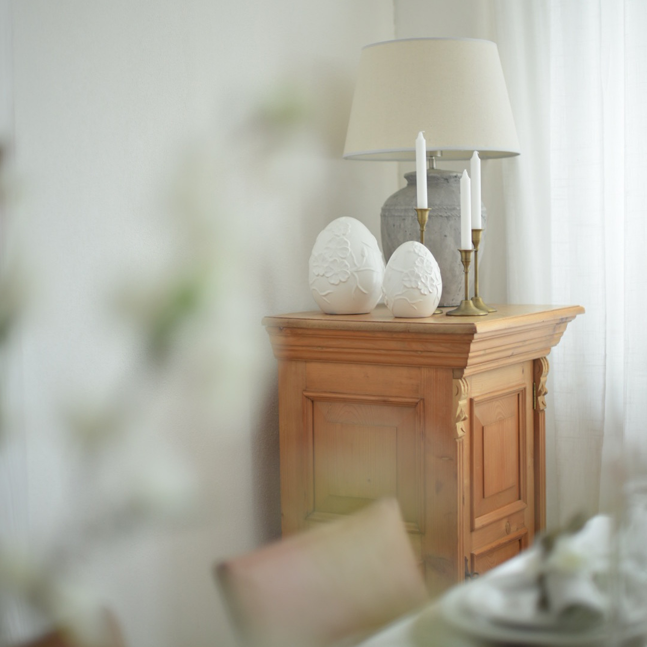 Dřevěná skříňka s lampou, svíčkami a bílými keramickými dekoracemi ve tvaru vajec v světlém interiéru