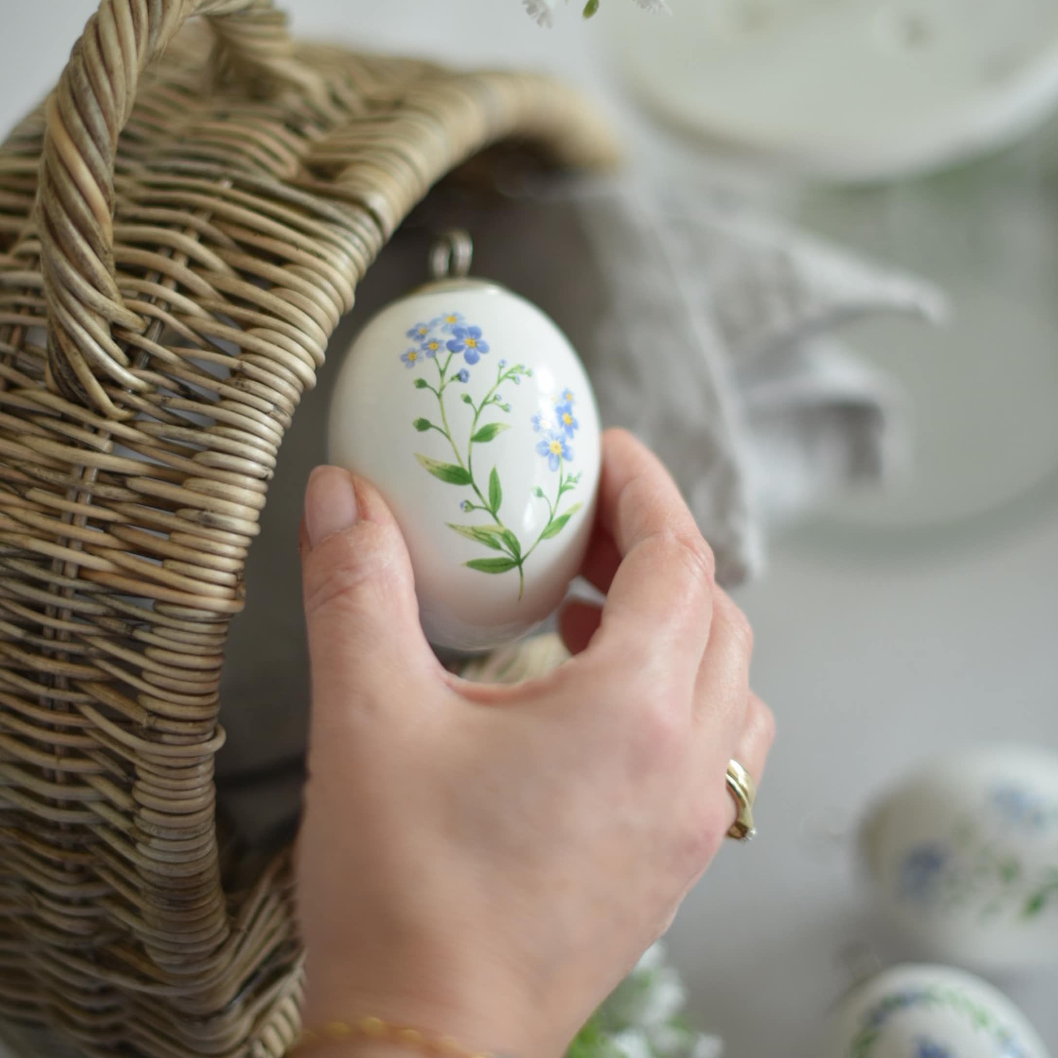 Ruka držící bílou keramickou velikonoční ozdobu s modrými květy vedle proutěného košíku