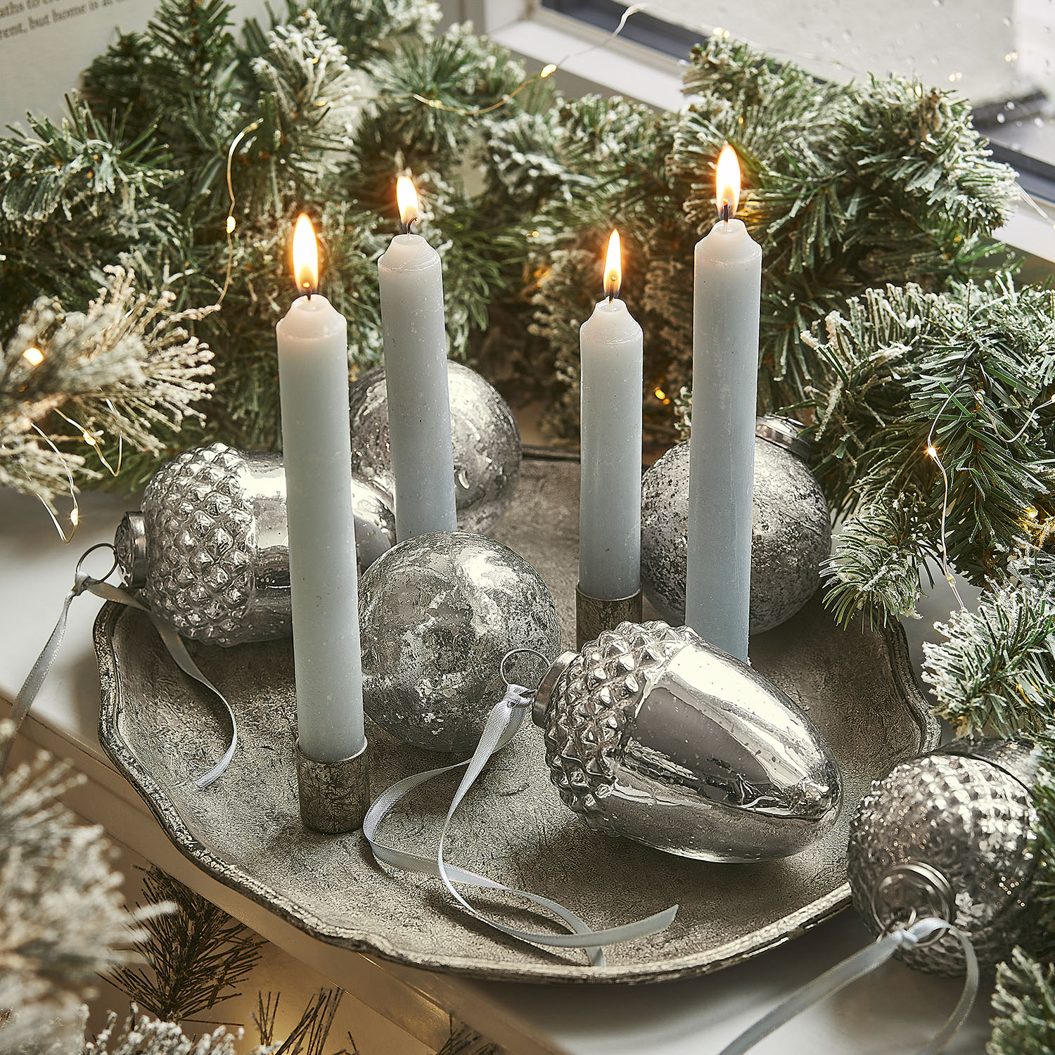 Svícen se čtyřmi modrými svíčkami a stříbrnými ozdobami na kovovém podnosu s zelenými větvičkami